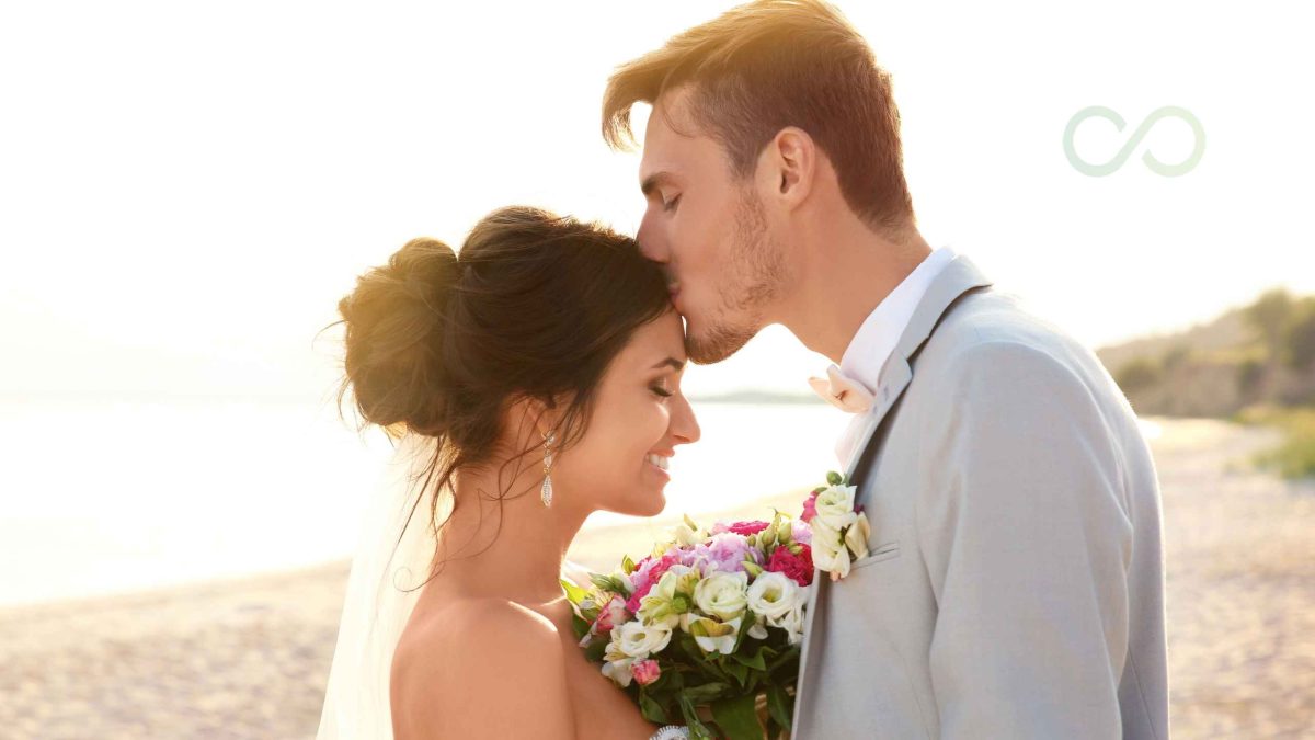 A newlywed couple sharing a tender moment on the beach, with the groom kissing the bride on the forehead as she holds a colorful bouquet of flowers in the warm sunset light Marketing to eco-conscious couples without greenwashing Green Loop